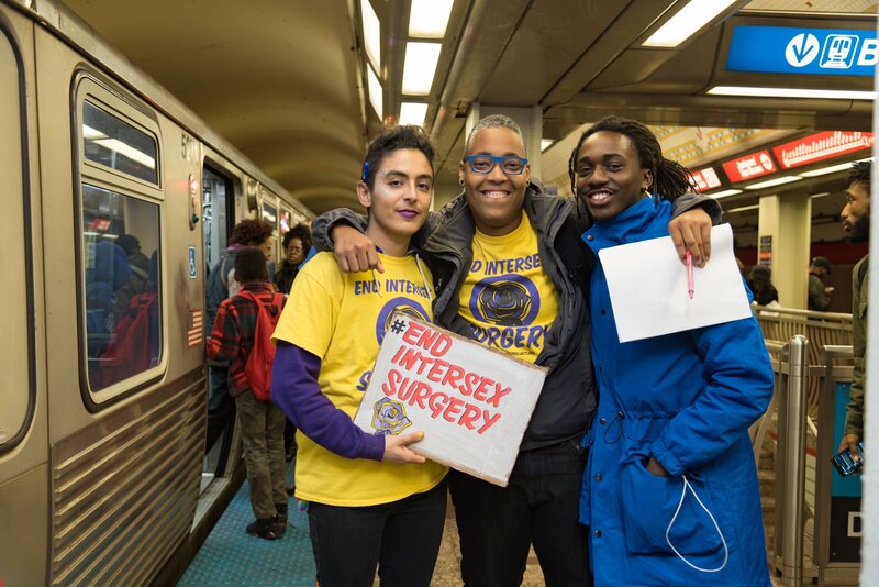 Saifa smiles inside a train station. He has his arms hanging over and hugging Pidgeon and Ric. Pidgeon holds an EndIntersexSurgery sign.