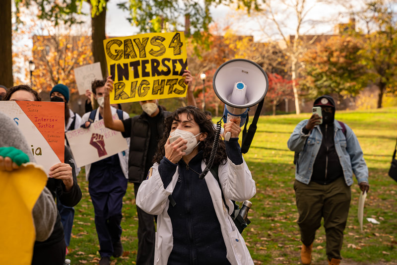 A medical student in a white coat holds a megaphone at an intersex justice rally outside Columbia in New York. In the background another student holds a sign, "Gays 4 intersex rights." All wear masks for covid safety.