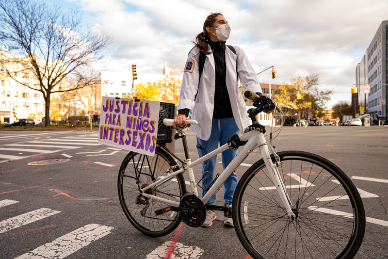 A medical student wears a white coat and stands for a hero-style portrait holding a bicycle. The student looks away into the distance. Taped onto the bike is a sign in Spanish that reads "Justicia para ninos intersexos."
