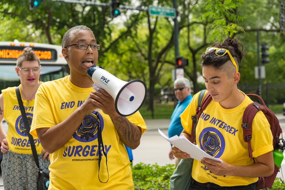 Saifa, IJP's co-founder, a Black intersex man with short gray hair, speaks through a megaphone at a protest. Standing next to him is Pidgeon, IJP's other co-founder, a nonbinary person with light brown skin and a mohawk haircut, reading from a piece of paper.