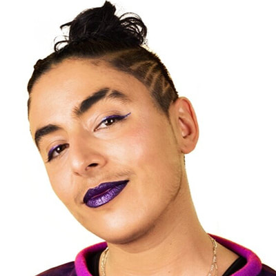 Pidgeon, an intersex activist, looks up confidently at the camera. They have light brown skin and dark hair pulled up into a bun. The side of their head's undercut is shaved in an undercut pattern. They have on purple lipstick and eyeliner.
