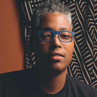 Sean Saifa Wall, a Black intersex man, in glasses and a t-shirt standing against a patterned wall with soft, dark lighting.