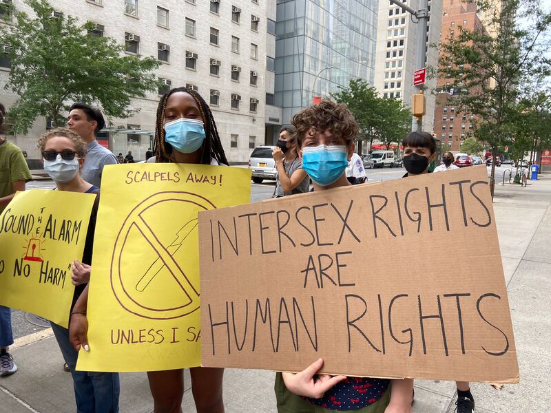 Students and doctors stand on the sidewalk in masks holding signs, "Scalpels away unless I say," and "Intersex Rights are Human Rights."